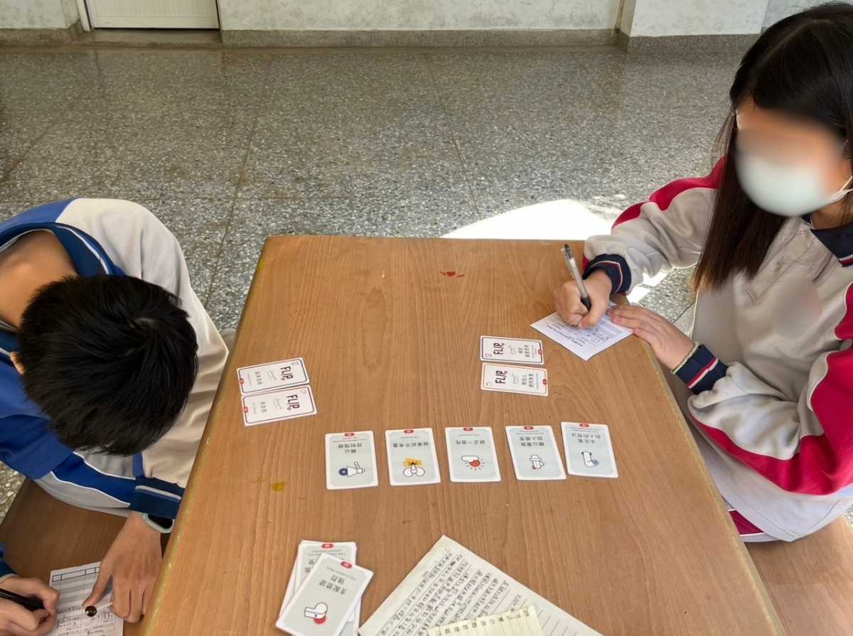 Two students sit at a classroom table and play a personality card activity. Several cards are arranged in the center of the table while the students write their choices on recording sheets.